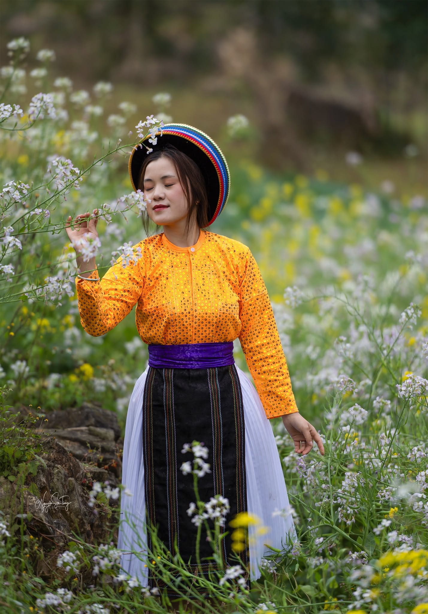 Thiếu nữ áo cam đội mũ thổ cẩm giữa vườn hoa trắng
