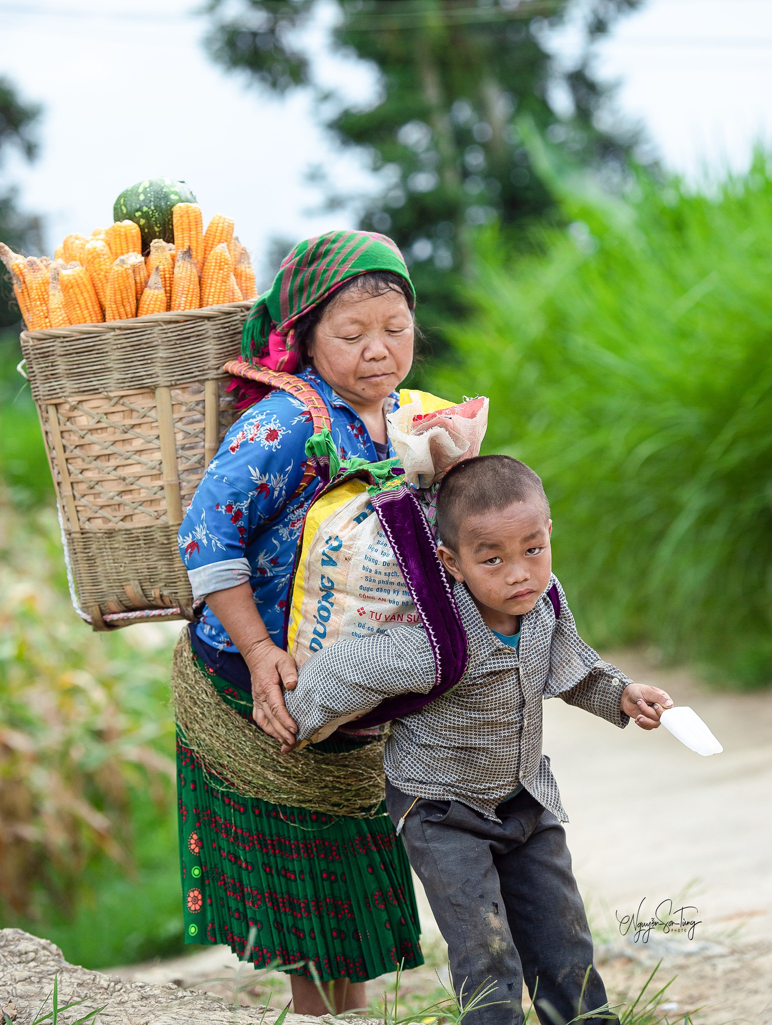 Người mẹ gùi ngô, dắt tay con trai đi trên đường