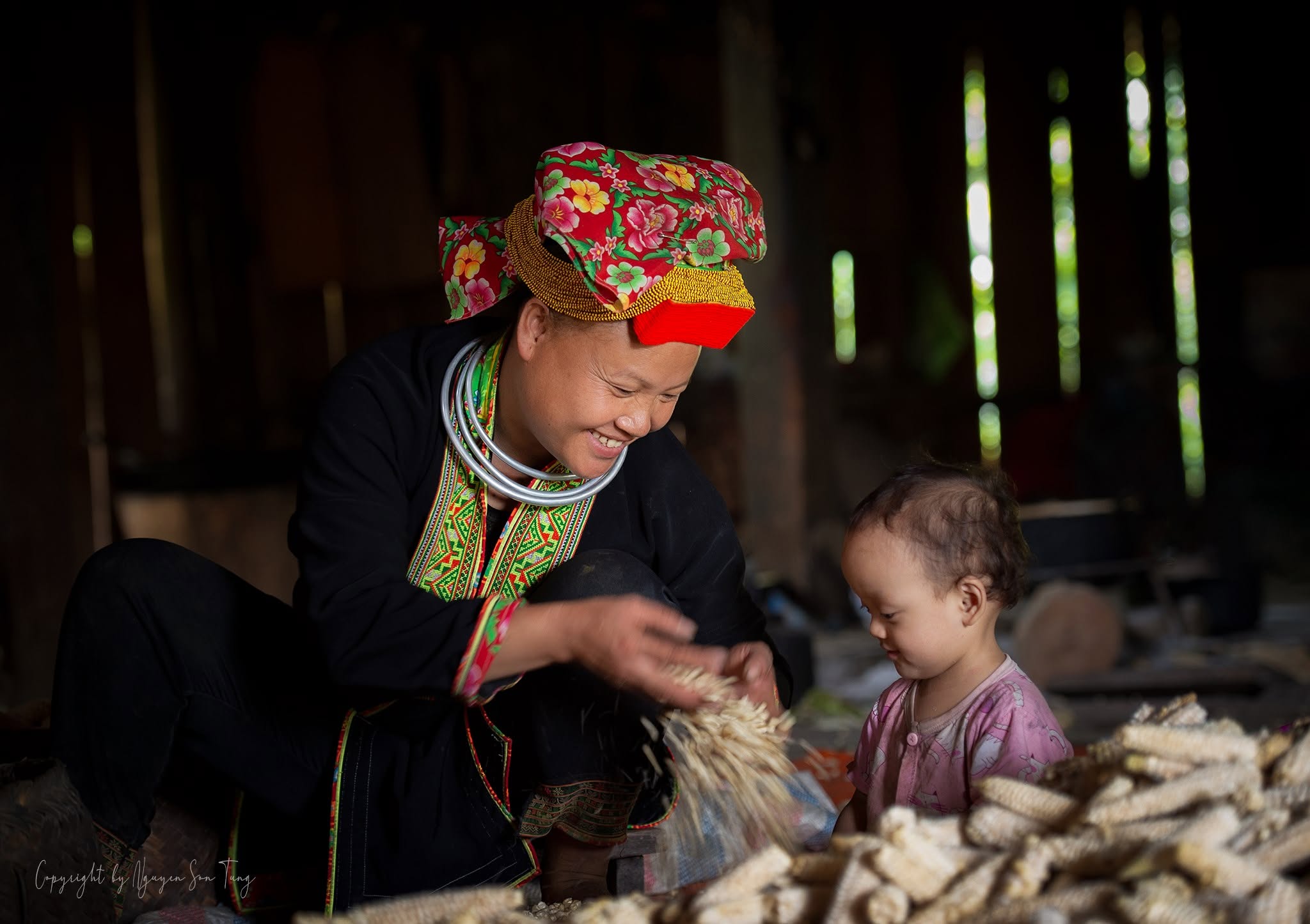 Người phụ nữ Dao sàng thóc ngô trong nhà cùng đứa cháu nhỏ