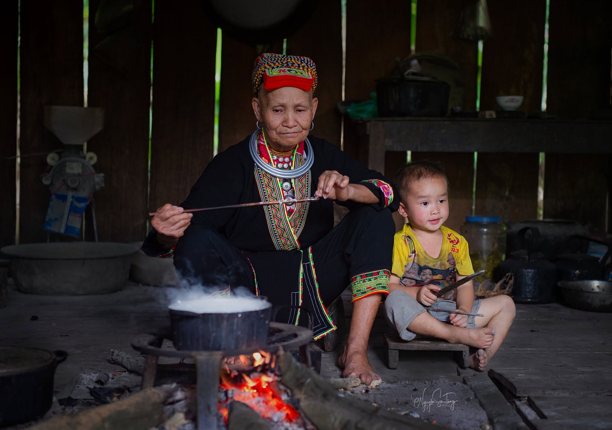 Cụ bà Dao ngồi bên bếp lửa giữa nhà cùng đứa cháu nhỏ